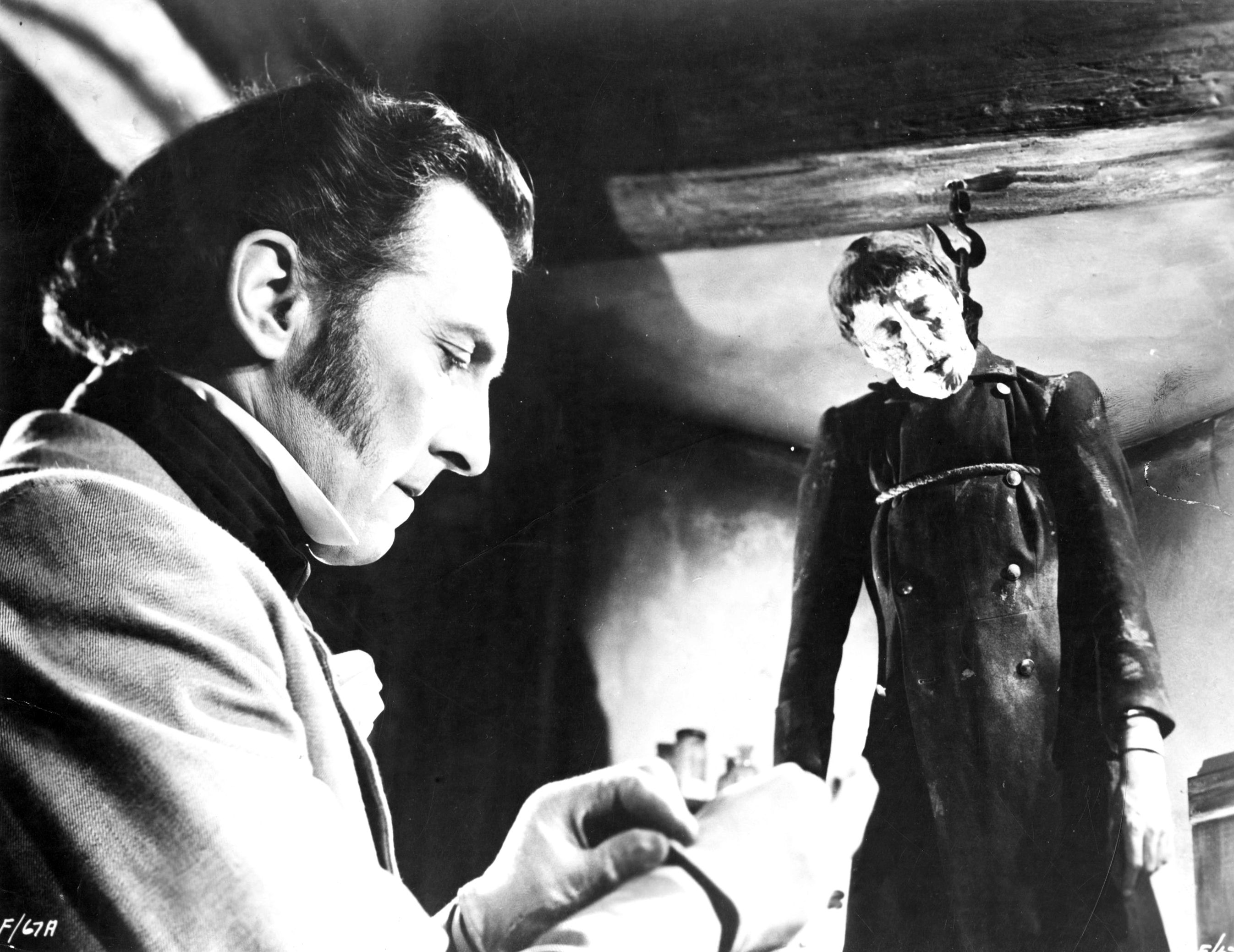 A ruthlessly amoral Baron Frankenstein (Peter Cushing) (l) prepares while his monster (Christopher Lee) (r) hangs on a hook in The Curse of Frankenstein (1957)