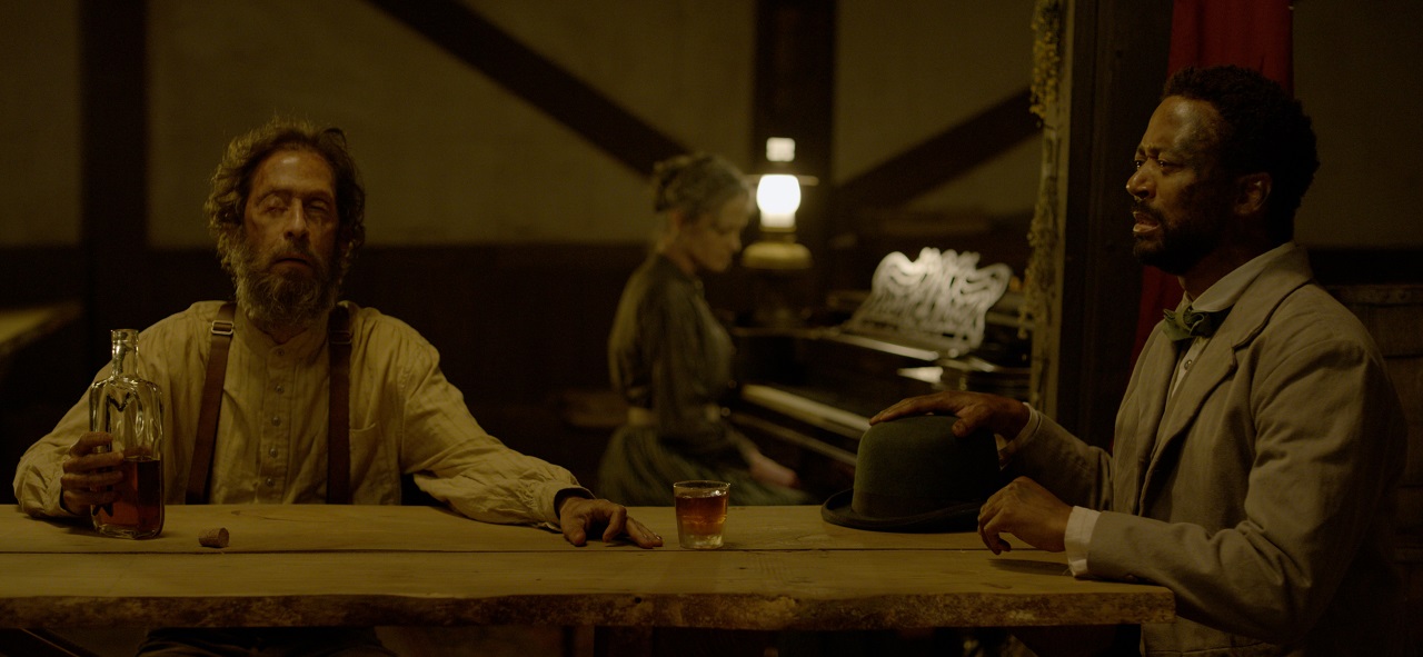 Blind barman Torb (Tim Blake Nelson) and newcomer James McCune (Thomas Hobson) with Angela Bettis at the piano in the background in Ghosts of the Ozarks (2021)