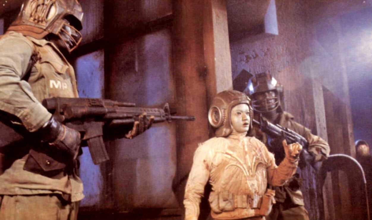 The android Grid (Deep Roy) surrounded by military droids in Starship (1984)