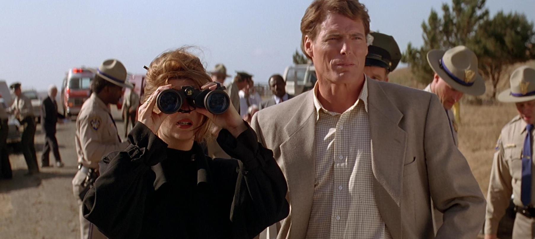 Christopher Reeve and Kirstie Alley (behind binoculars) watch from the military cordon line in Village of the Damned (1995)